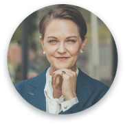 The image shows a professional headshot of a woman in a formal outfit, with her hand placed thoughtfully near her chin. She has short hair, is wearing pearls, and is giving a confident, slightly contemplative smile. The background is blurred, with a soft outdoor setting, and the image is framed in a circular shape.