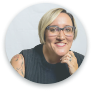 A circular headshot of a woman with short blonde hair, glasses, and a tattoo on her arm. She is smiling and resting her chin on her hand. The background is light-colored.