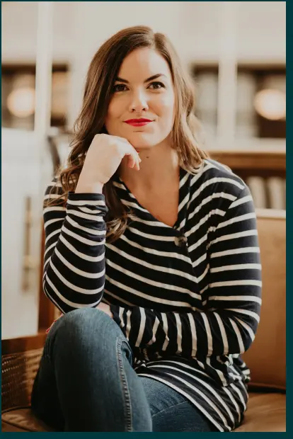 The image features a woman with long brown hair, wearing a striped, navy and white shirt. She is sitting comfortably with one hand resting on her chin and a thoughtful expression. Her makeup is natural, and she appears to be in a well-lit, cozy indoor setting.