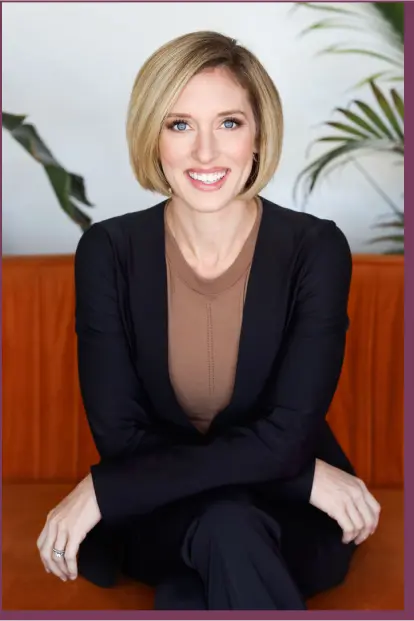 The image shows a woman with short blonde hair sitting on a modern orange couch. She is dressed in a neutral brown top and a black cardigan, smiling confidently at the camera while crossing her arms. Behind her are green plants, contributing to a relaxed and welcoming atmosphere.