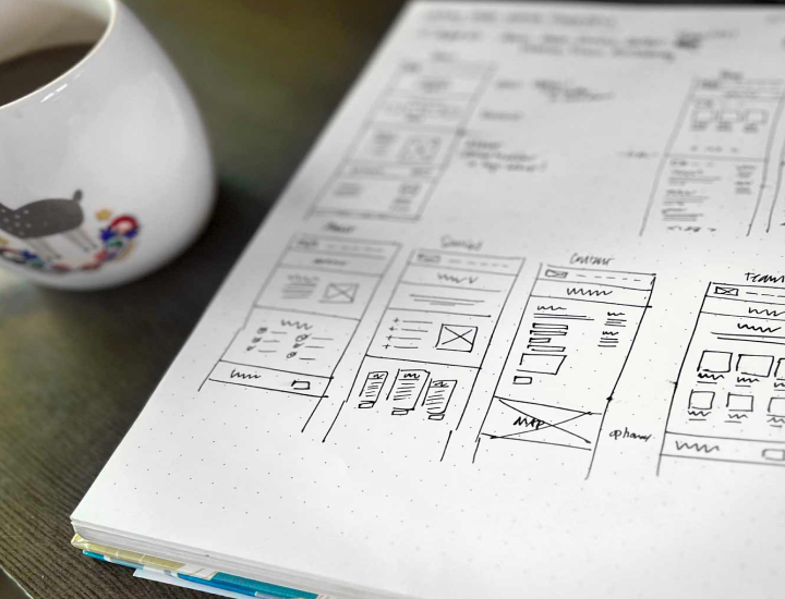 The image features a close-up of a notepad with hand-drawn wireframe sketches for a website design. The sketches display various page layouts, including sections like "About," "Contact," "Services," and "Team." A coffee mug is visible in the foreground, with a playful design on it, while the notepad is placed on a dark wood surface.