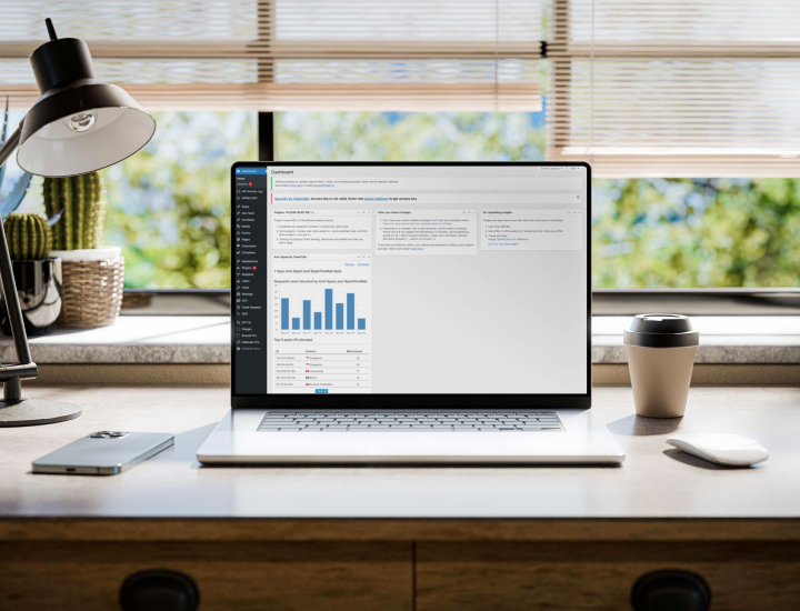 The image displays a laptop on a desk, showing a digital dashboard interface on the screen. The workspace features a lamp, a potted plant, a smartphone, and a coffee cup, with a bright, natural light filtering through the blinds.