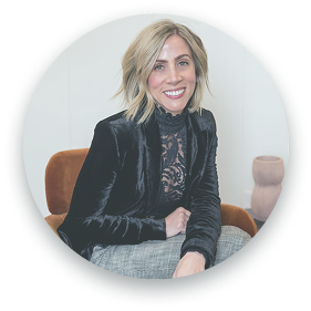 The image shows a professional portrait of a woman with short blonde hair, wearing a black velvet blazer over a black lace top. She is sitting in a chair and smiling warmly at the camera. The photo is framed in a circular crop, with a neutral background and soft lighting.