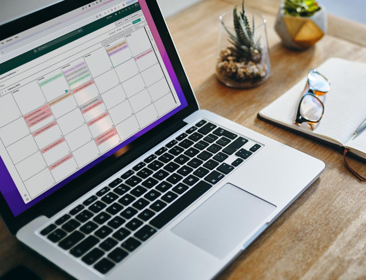 The image displays a laptop open on a desk with a visible calendar application on the screen. The calendar has various events and tasks color-coded, with some entries marked in red. On the desk, there is also a notebook, a pair of glasses, and a small potted plant, contributing to a neat and organized workspace. The laptop's keyboard and screen show that the person is likely working on scheduling or managing their tasks.