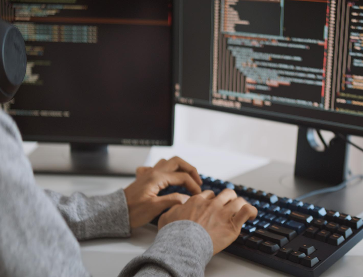 The image shows a person typing on a mechanical keyboard in front of two computer monitors displaying lines of code. The individual is focused on the task at hand, possibly engaged in software development or programming, with the monitors featuring dark backgrounds and colorful code snippets. The setting suggests a work environment designed for coding or technical tasks.