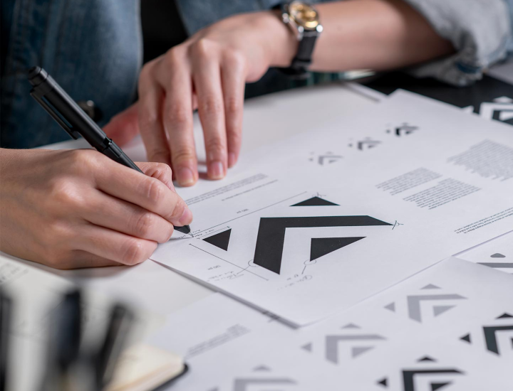 The image shows a person drawing or refining a logo design on paper. The design appears to be a geometric pattern, featuring bold, black triangular shapes arranged in a dynamic, angular formation. The person is using a pen to add precise measurements or adjustments to the drawing, while several iterations of the logo are scattered across the table. The workspace is neatly arranged with some office supplies visible in the background.