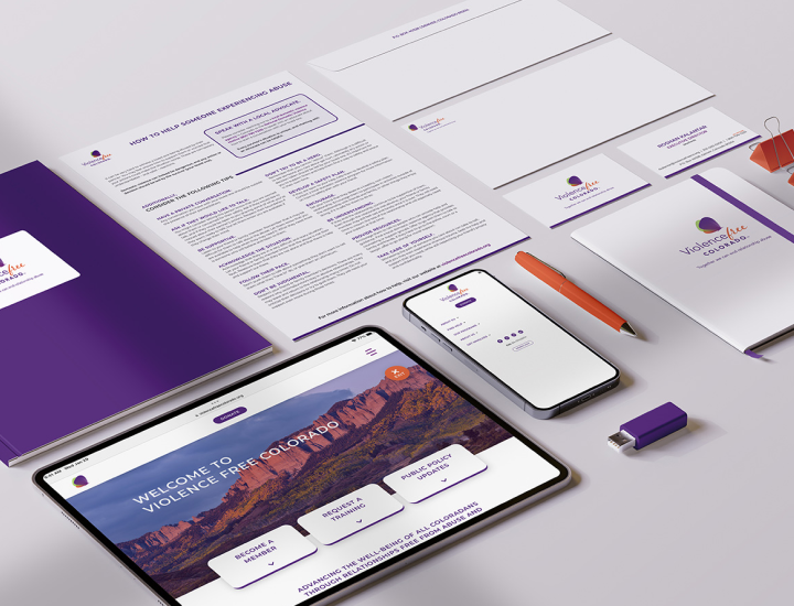 The image displays a collection of branded materials for an organization called "Violence Free Colorado." The set includes a variety of office supplies, such as a notebook, pens, letterhead, business cards, and a USB drive, all in a cohesive purple and white color scheme that matches the brand. The materials are arranged alongside digital devices displaying the website for Violence Free Colorado, which features a beautiful landscape photo as the website background. The brand's modern and professional appearance is highlighted by the tablet and smartphone showcasing the organization's online presence.